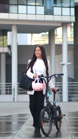 vertical Confident multicultural business woman walking with her sustainable electric folding bike in a modern financial district, choosing an eco-friendly way of commuting to her office