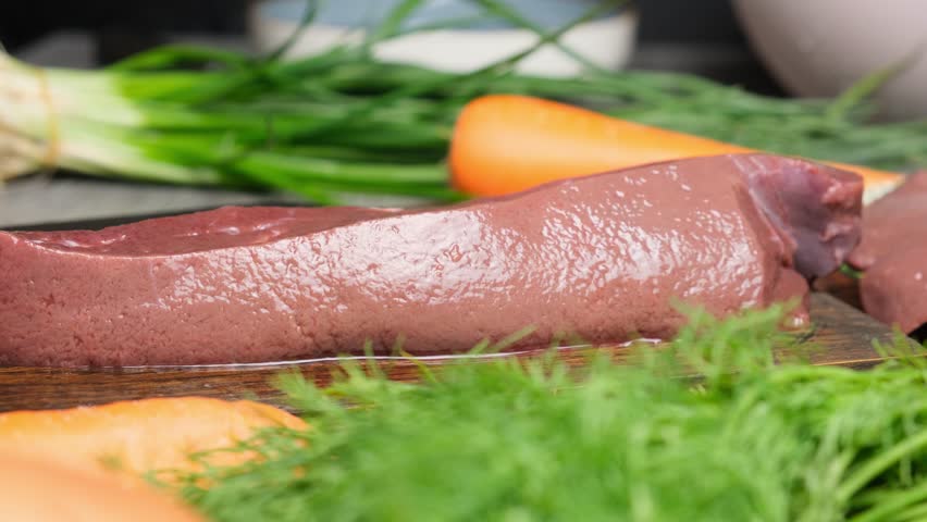 The camera moves close to a piece of fresh liver shining in the light on a wooden cutting board among vegetables and herbs. Raw beef liver before cooking. Juicy liver in a restaurant kitchen.