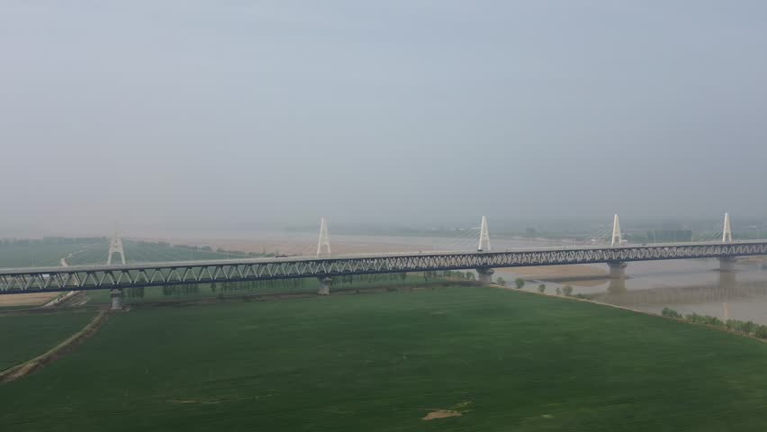 Aerial of Zhengxin Yellow River Rail-Road Bridge, Zhengzhou, facilitating road-rail traffic across Yellow River at spring noon, with green fields and mist.