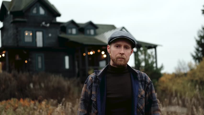 Confident man in plaid jacket stands in a field with a rustic house behind him, showcasing his expression and the ambiance, camera zooms in for detail