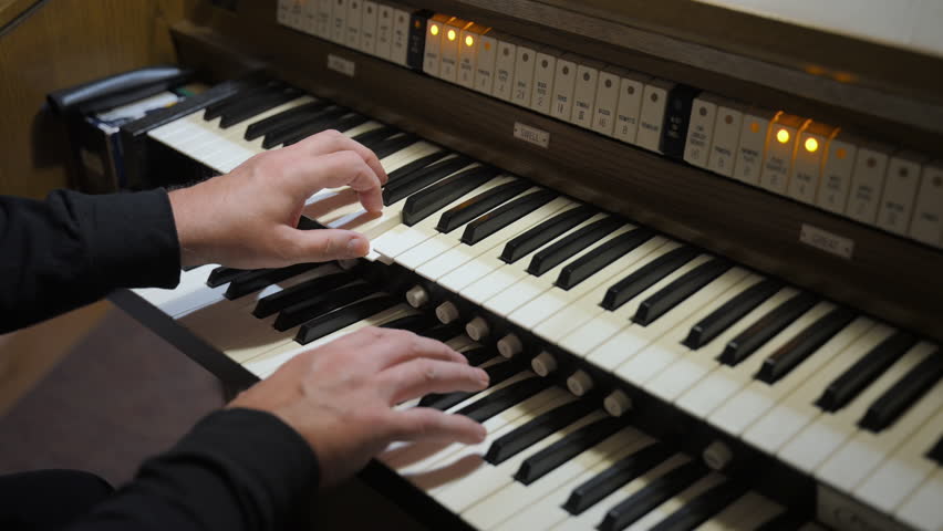 Musician plays an organ inside a church or concert hall. The performer hands are actively moving across multiple manuals keyboards, suggesting live performance. 