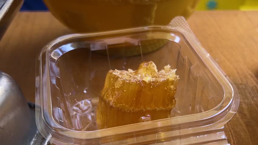 Horizontal video. Honey seller places piece of juicy honeycomb by knife into plastic container on scale at New Year farm fair, Christmas market, autumn harvest festival Tbilisoba, city holiday