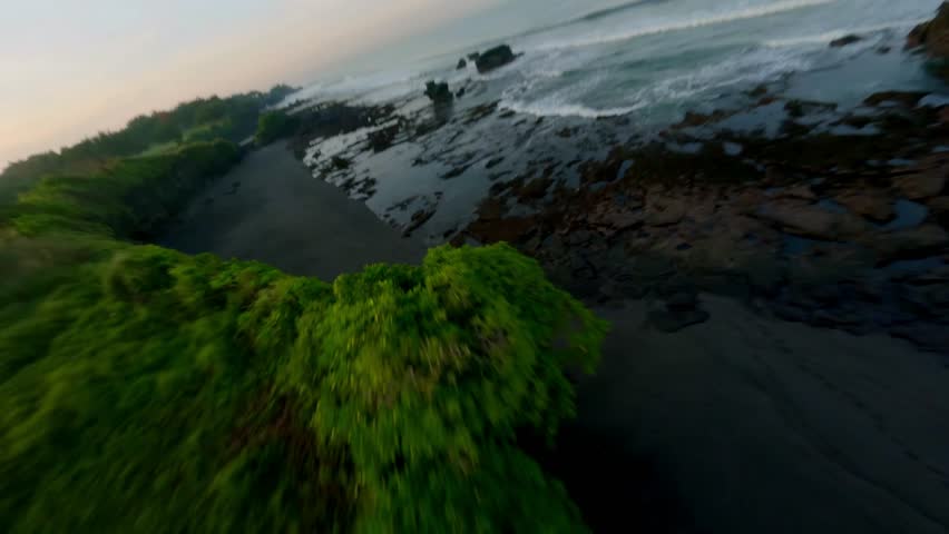 Atmospheric FPV drone footage flying fast and low over a rocky coastline in Bali during a hazy sunrise. The camera glides over the dark volcanic sand and moss-covered rocks in the shallow water.