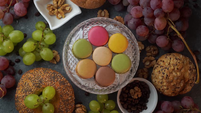 A seamless overhead loop shows assorted macarons on a glass plate surrounded by red grapes, rustic buns, coffee beans, and walnuts on a gray stone tabletop.