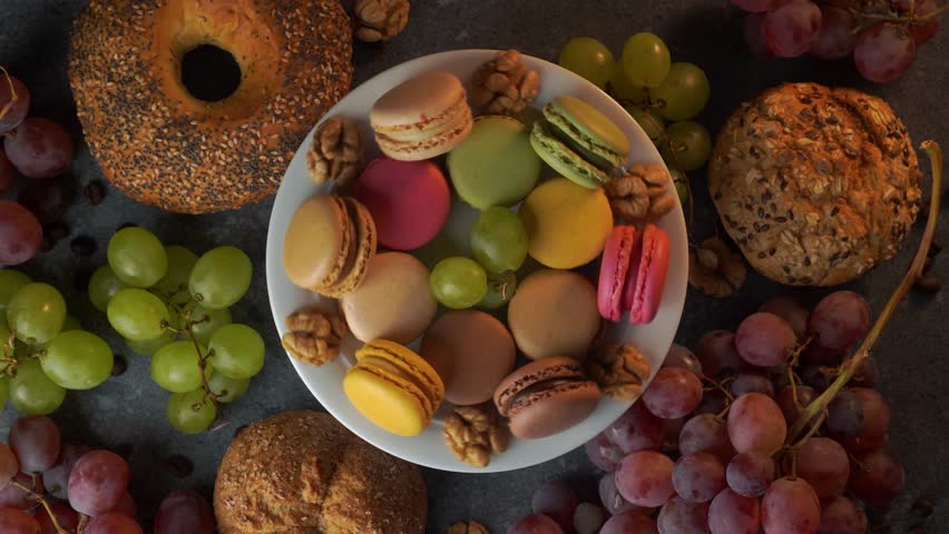 A colorful flatlay loop presents macarons in a white bowl with red grapes, buns, coffee beans, and walnuts. The plate rotates on a gray stone tabletop to create a dynamic edible background