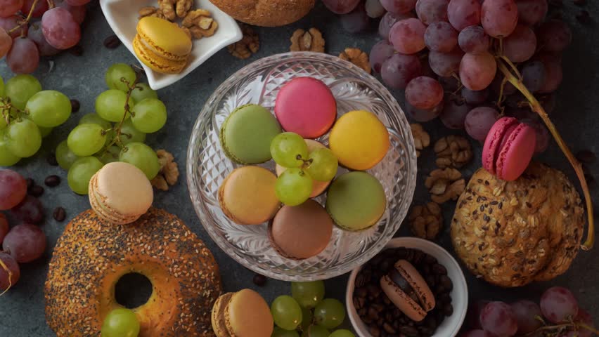 A colorful top down loop shows macarons arranged on a glass plate with red grapes, buns, coffee beans, and walnuts. The composition spins smoothly on a gray stone tabletop for titles or branding.