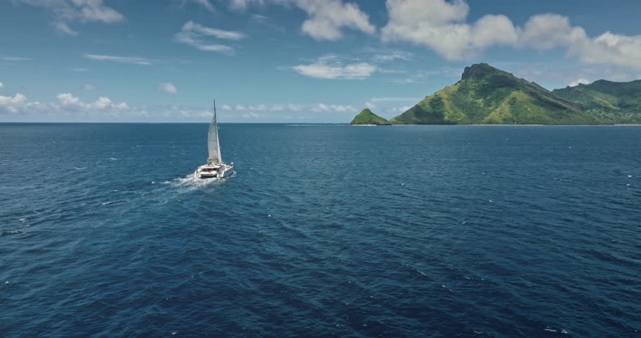 Fiji: Catamaran sailing blue ocean near tropical island with lush vegetation and turquoise water, perfect travel destination for summer vacation. Remote wild nature paradise. Drone flight footage