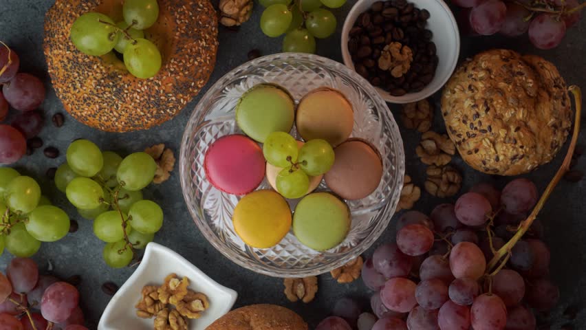 A clean looped flatlay features a glass plate of colorful macarons framed by red grapes, bakery buns, coffee beans, and walnuts. Smooth rotation over gray stone provides a ready to use food background