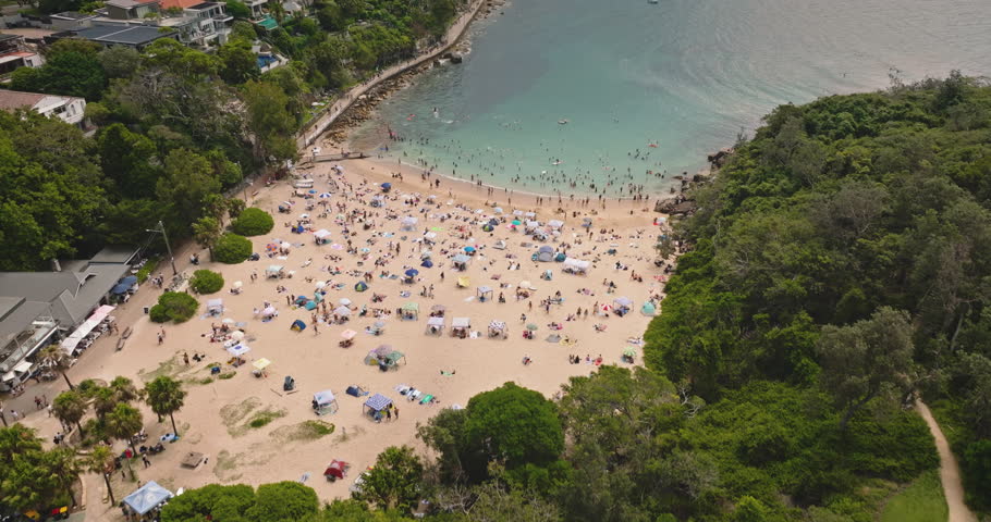 Manly Beach, Sydney, Australia: People enjoying clear turquoise water and sandy shoreline, crowd swimming, sunbathing, and relaxing under umbrellas on sunny summer day. Modern cityscape in background