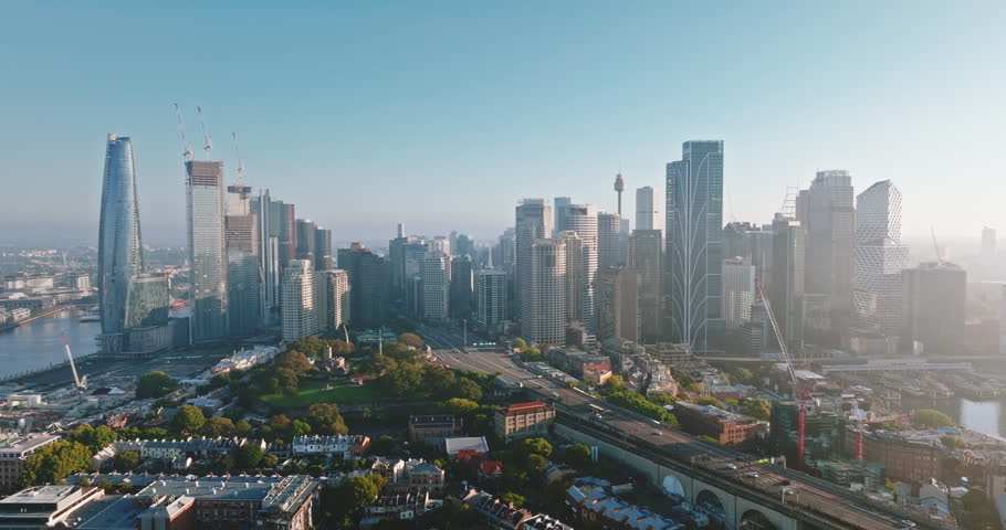 Sydney city skyline with modern Barangaroo high-rise buildings skyscrapers, road, historic wharves, and iconic Sydney Harbor Bridge spanning the vibrant blue harbor water. Aerial zoom out panorama