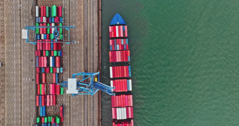 Cargo ship docks at a busy industrial port, having colorful shipping containers loaded and unloaded by cranes, representing global logistics and transportation. Top down aerial drone flight