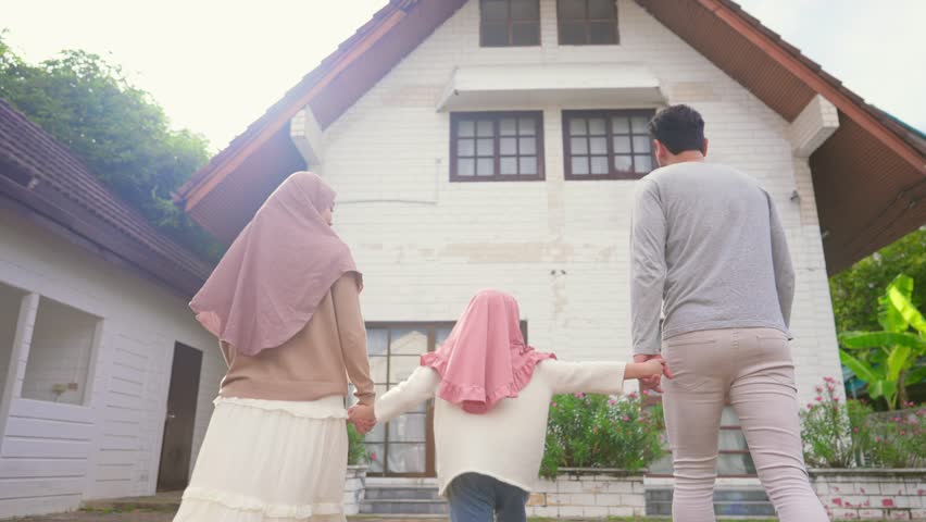 Happy Muslim family holding hands and looking proudly at their new house. Attractive couple standing together outdoors near home entrance, symbolizing love and celebrating new real estate ownership.