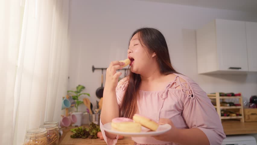 Asian plus-size woman enjoy eating unhealthy snack in kitchen at home. Attractive smiling overweight curvy girl feel happy and relax, bites a sweet donut on cooking counter for breakfast in cozy house