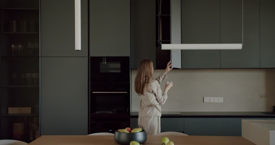 Woman in pajamas walks through her modern kitchen and opens a cupboard. The kitchen is decorated in a minimalist style with dark gray cabinets and a light wood table. She holds a glass of water.