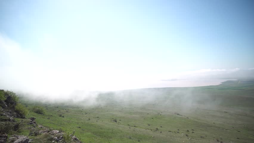 A foggy hillside with a clear blue sky in the background. The sky is dotted with clouds, and the grass is lush and green