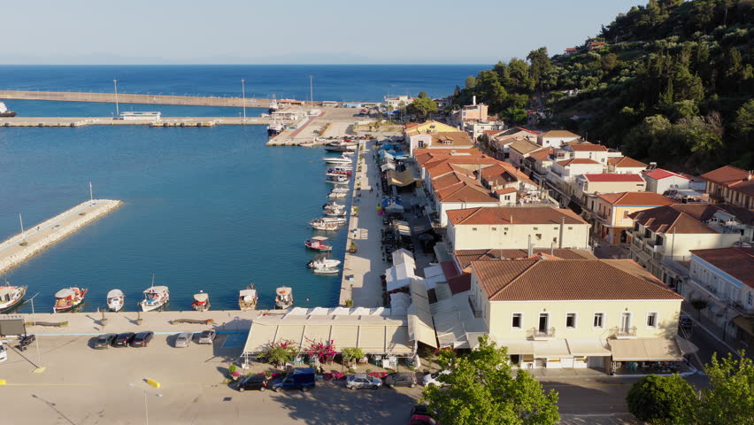 Aerial shot flying above the seaside promenade of Katakolo port, Greece, with cafes lining the waterfront and fishing boats anchored in the calm bay