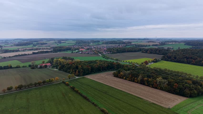 Aerial establishing shot of colored farmland fields and forest trees. Small american village in background during cloudy day in October. Wide shot. Peaceful rural nature landscape in US