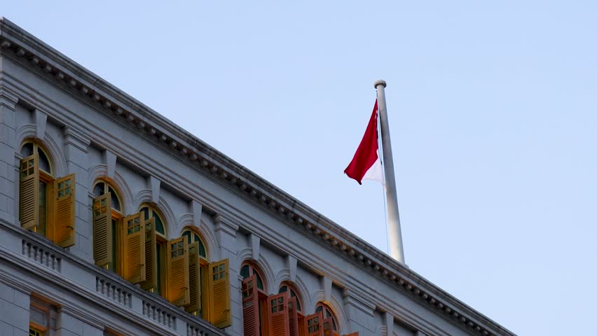 Singapore flag waves atop ornate building with colorful window shutters, captured in steady daylight shot