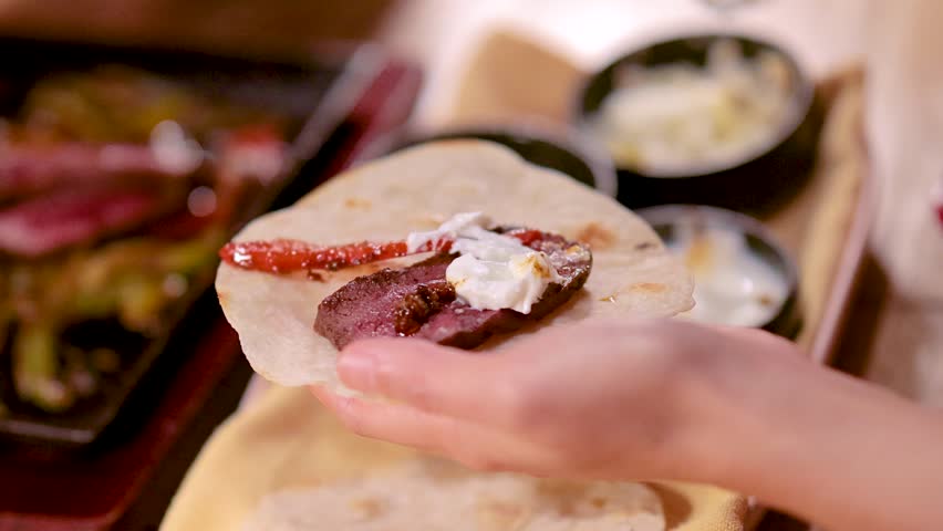 Hand adds cheese to beef taco with salsa and sour cream, warm indoor lighting, close-up
