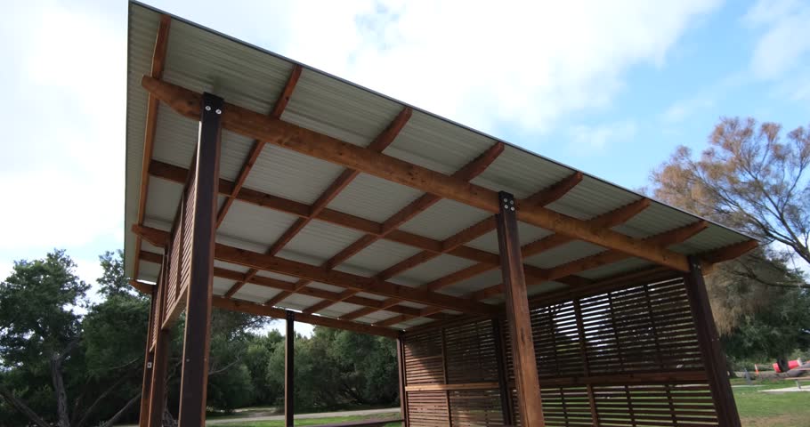 A wooden picnic shelter with a slatted privacy wall, tables, benches, and a corrugated metal roof stands in a public park. Offering shade and seating for families or groups to enjoy outdoor gathering