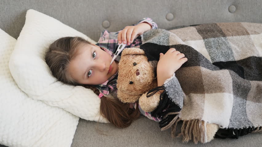 A young girl, wrapped in a blanket, rests on a couch while taking her temperature with a thermometer, hugging a teddy bear.