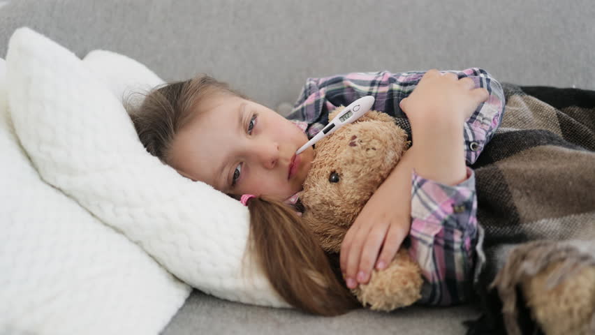 A young girl lies on a couch, resting with a teddy bear and a thermometer in her mouth, indicating illness.