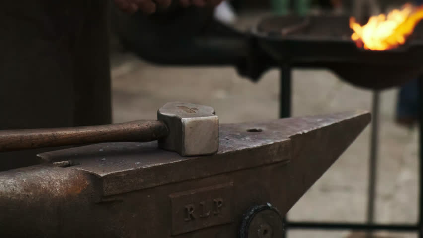 Static close-up of a blacksmith striking red-hot iron on an anvil with a hammer; the coal forge blazes softly out of focus in the background.
