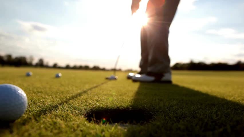 Golfer stands on putting green at sunset with multiple golf balls nearby, concentrating on sank shot, casting long shadows across vibrant grass.