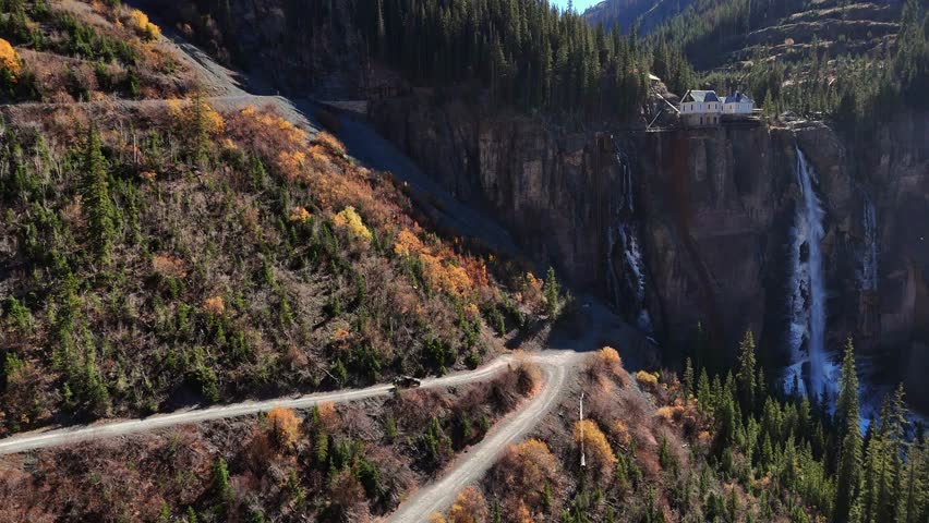 Aerial view of Bridal Veil Falls cascading down the cliff face, framed by autumnal foliage and a winding dirt road, Telluride, Colorado, United States.