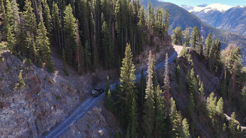Aerial view of a winding mountain road cutting through dense forests against a backdrop of snow-capped peaks, Telluride, Colorado, United States.