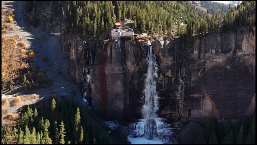 Aerial view of Bridal Veil Falls cascading down rugged cliffs, contrasting with the verdant trees and the buildings on the clifftop, Telluride, Colorado, United States.