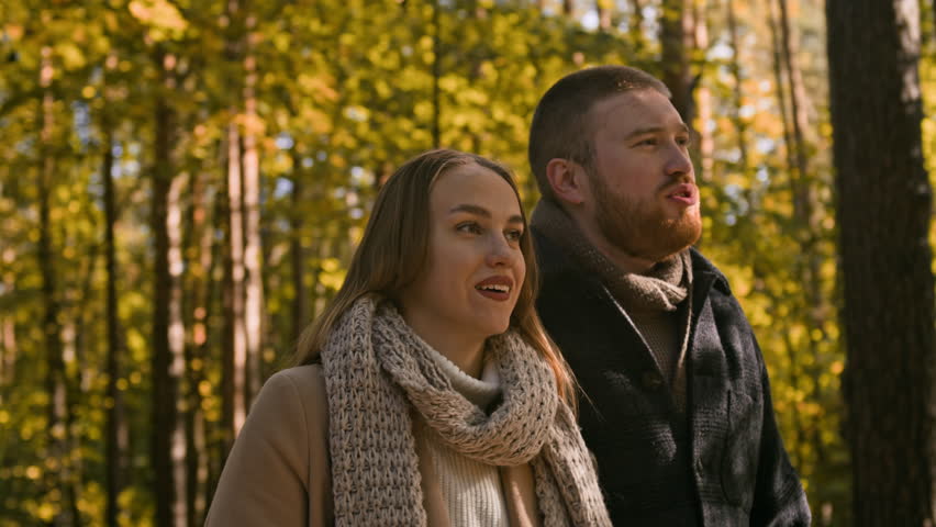 Medium tracking shot of loving Caucasian couple enjoying relaxing walk in autumn park with trees and multicolor foliage in morning