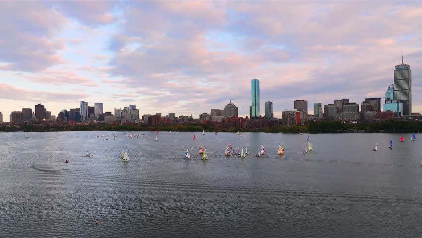Aerial view of sailboats gliding on the Charles River with the Boston skyline of skyscrapers and urban parks, under a dramatic sunset. Massachusetts, USA.