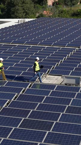 Three engineers wearing hard hats and safety vests walking across a rooftop covered with photovoltaic solar panels while inspecting the installation and checking data on a laptop and tablet
