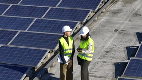 Professional female engineers wearing hard hats and safety vests discussing a project on a rooftop covered with solar panels, one holding a clipboard and the other using a digital tablet - Powered by Shutterstock - Get 15% off with code: PIKWIZARD15