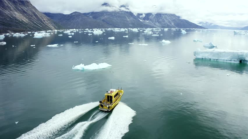 Aerial view of a yellow boat sailing across the water, contrasting with white wakes and floating icebergs, near mountains, Ilulissat, Avannaata, Greenland.