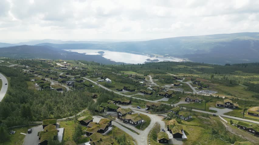 Aerial view of Beitostolen village with dark cabins and green roofs, nestled among lush trees near a winding road and a distant lake, Beitostolen, Innlandet, Norway.