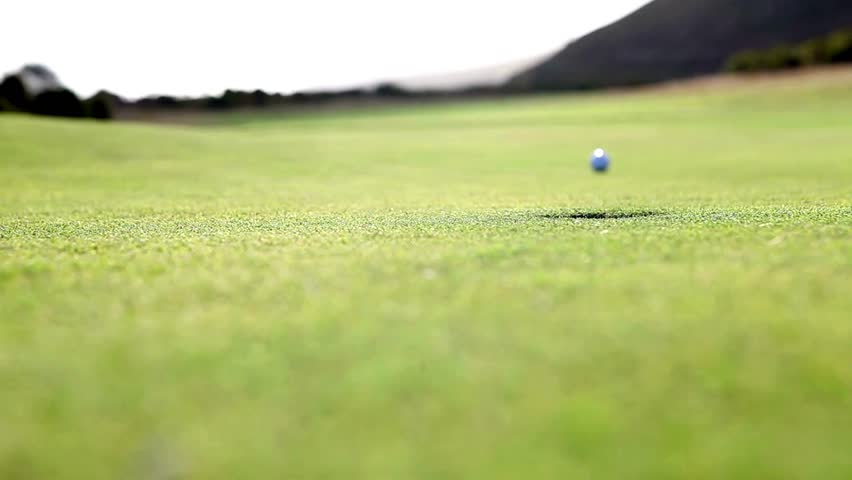 Golfer takes a shot on a beautiful green golf course. Ball rolls across well-maintained grass towards the hole under bright sunlight, showcasing skillful gameplay.