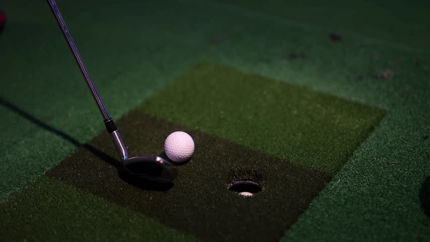 Golfer takes aim with club, ready to hit golf ball resting on artificial turf, targeting hole in indoor driving range environment.
