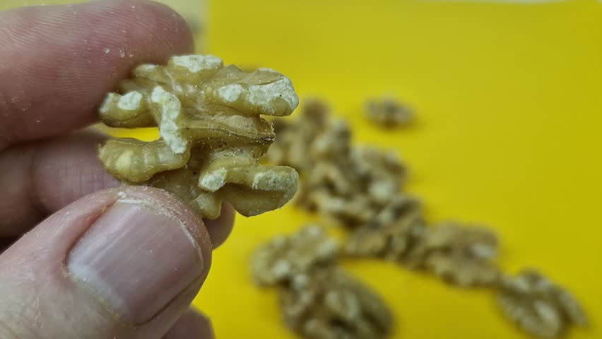Ultra-close 4K macro view of a single shelled walnut half being held between the thumb and forefinger of a person, set against a bright yellow background.