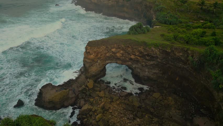 An aerial view of a wooded rugged Coastline with a natural Sea Arch and foamy waves at Karang Bolong Beach in Indonesia