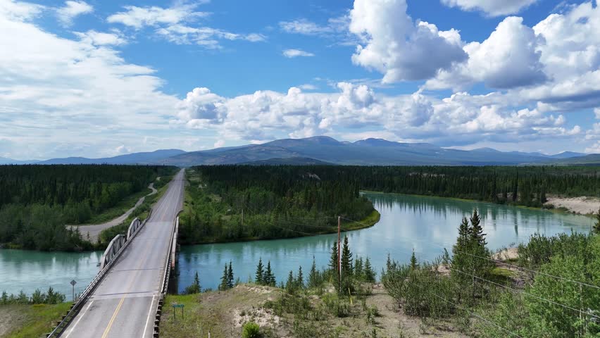 Vast natural landscape with a winding road crossing a turquoise river towards distant blue mountains under a dynamic cloudy sky in Alaska
