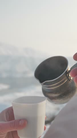 Vertical video, over-the-shoulder view of a woman holding a plastic cup and pouring hot coffee from a cezve against snowy mountains. Hot drink outdoors in the mountains during a hike.