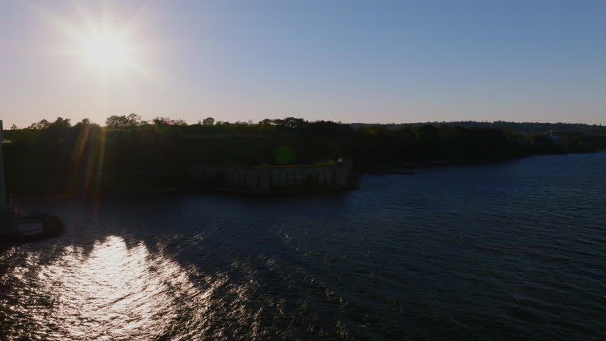 Aerial view of the Verrazzano-Narrows Bridge over Battery Weed fort in Staten Island, New York, with sunlight reflecting on the water and Manhattan skyline in the distance. ProRes HQ422.