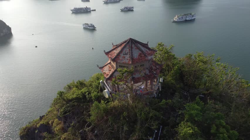 4K drone reveal shot pulls back from the iconic Ti Top Island pagoda to unveil a super-wide, panoramic vista of Ha Long Bay, dotted with numerous cruise ships and limestone karsts.