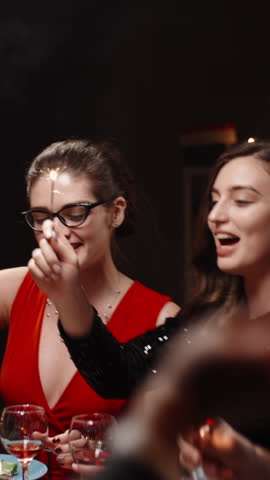 Vertical cinematic slow motion video joyful friends raising glasses with sparklers at festive dinner table cozy warm light holiday atmosphere concept friendship celebration christmas new year