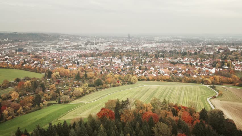 Camera glides back over vibrant autumn treetops, gradually revealing the cityscape of Ulm set beyond fields and woodland.