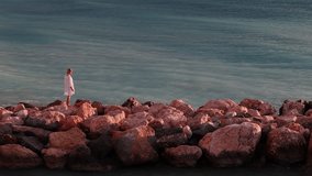 Middle-aged woman in white dress and hat stands alone on coastal rocks at sunrise, gazing calmly at the turquoise sea and enjoying a quiet peaceful morning. High quality 4k footage - Powered by Shutterstock - Get 15% off with code: PIKWIZARD15