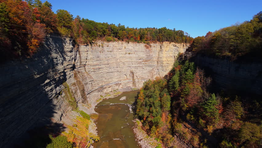 Drone view of the deep gorge and steep cliffs of Letchworth State Park, New York. The Genesee River winds between layered rock formations and colorful autumn forests, showcasing dramatic natural