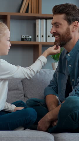 Happy Caucasian family at home daughter with father talking on couch cute child girl playing with loved dad tickling beard of daddy touching nose of lovely kid playful spending time together on sofa